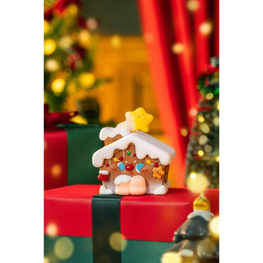Decorative gingerbread house figurine on a red and green box with Christmas tree and lights in the background.