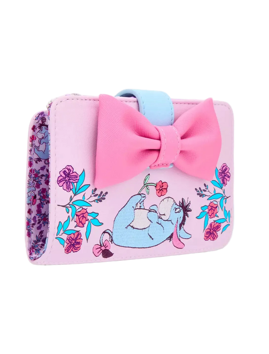 A pink faux-leather wallet featuring a large pink bow, a light blue strap, and an illustration of Eeyore sitting among colorful flowers. The side of the wallet shows a purple and pink floral pattern.