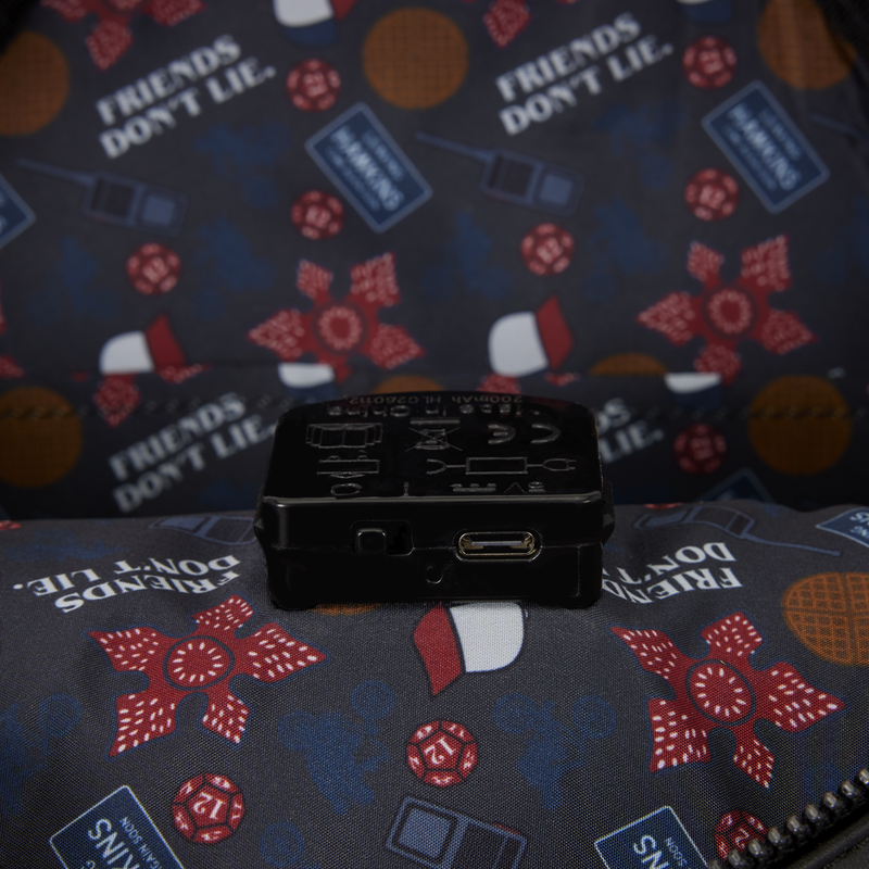 A small black electronic device with a USB-C port, placed on a patterned zippered pouch featuring poker-themed graphics and the text 'FRIENDS DON'T LIE'.
