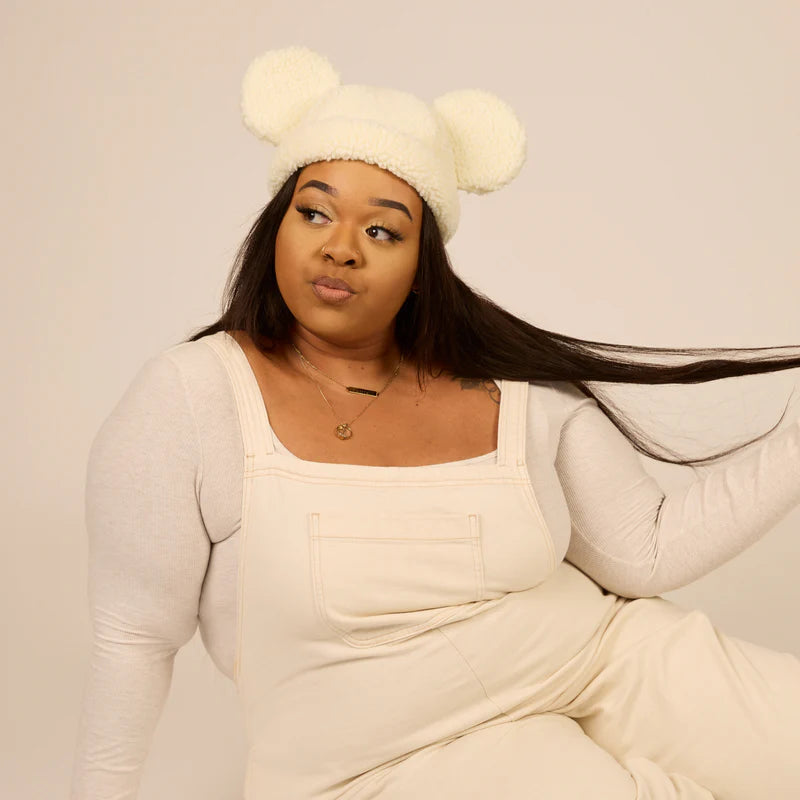 Person wearing a cream-colored outfit with a matching hat, sitting against a plain background
