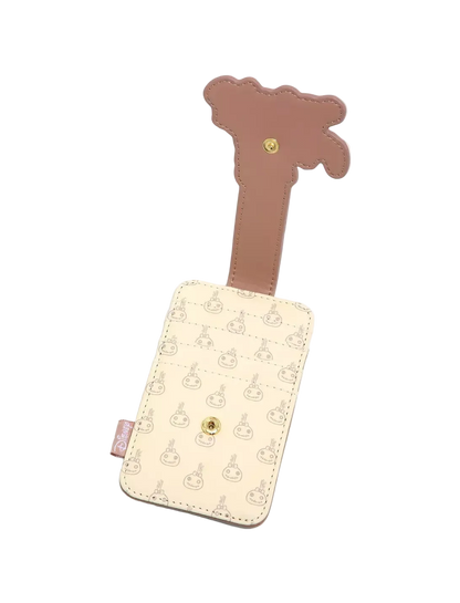 A cream-colored luggage tag featuring a repeating pattern of cartoon characters, with a brown strap shaped like a giraffe and a gold snap button closure.