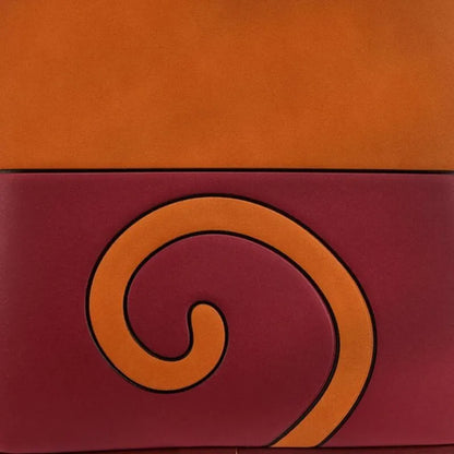 A close-up of a leather product, likely a wallet or clutch, featuring a two-tone design with a solid orange top section and a maroon bottom section. The maroon section is decorated with a large, raised orange spiral motif outlined in black.