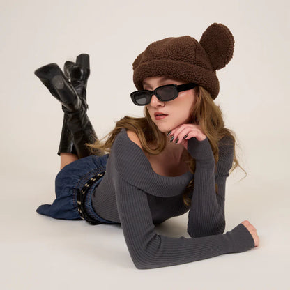 Woman wearing a brown knit hat with bear ears, black sunglasses, and black boots on a light background