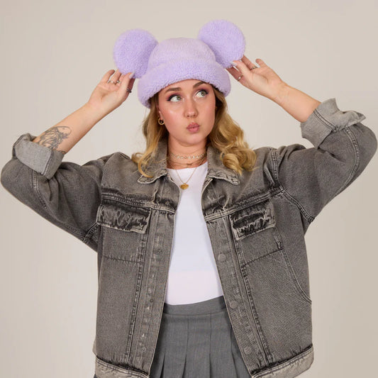 Person wearing a fluffy purple hat with large pom-poms, gray denim jacket, and gray pants on a plain background.