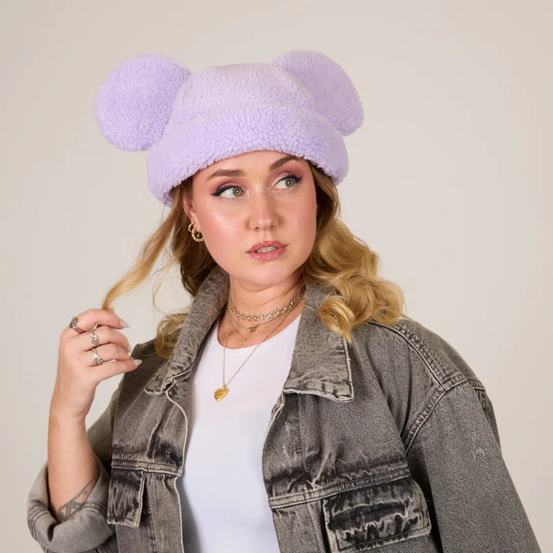 Woman wearing a lavender knit hat with large pom-poms against a plain background