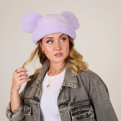 Woman wearing a lavender knit hat with large pom-poms against a plain background