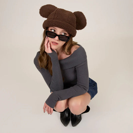 Woman wearing a brown teddy bear hat and black sunglasses on a plain background