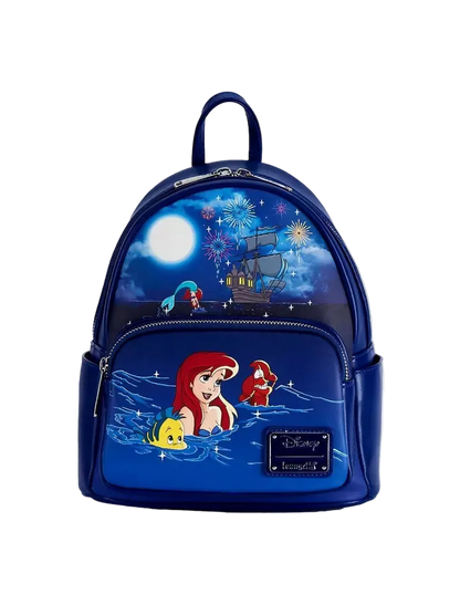 A Disney The Little Mermaid mini backpack in navy blue, featuring character artwork of Ariel, Sebastian, and Flounder under a starry night sky with fireworks and a ship, on a light gray background.