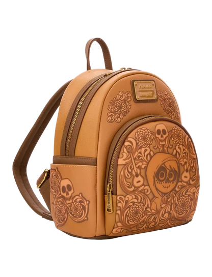 A tan faux-leather mini backpack with skull and floral tole-style embossing, featuring a top handle, adjustable shoulder strap, and gold-tone zipper pulls, shown on a light gray background.