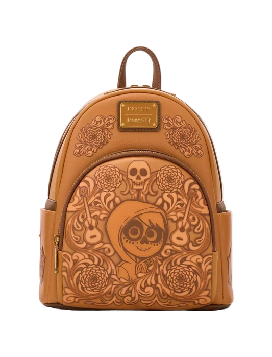 A tan leather mini backpack featuring an embossed Day of the Dead design with a central skeleton face, skulls, guitars, and floral patterns. The backpack has a top handle, zippered main compartment, side pockets, and a gold-tone metal brand plaque on the front.