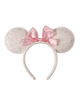 A Minnie Mouse-inspired headband with round ears covered in a light beige and pink Disney-themed pattern, featuring a large pink polka-dot bow on a white background.