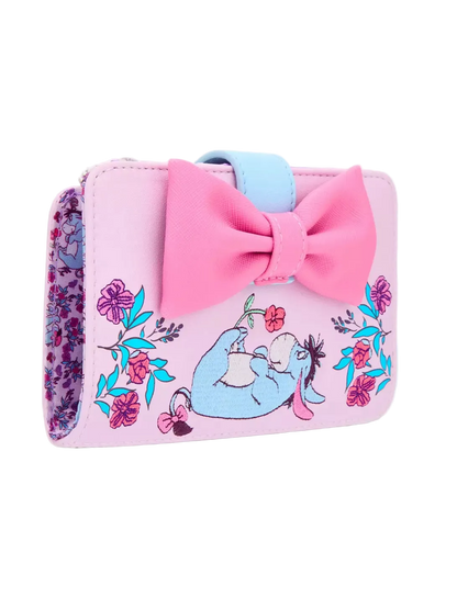 A pink faux-leather wallet featuring a large pink bow, a light blue strap, and an illustration of Eeyore sitting among colorful flowers. The side of the wallet shows a purple and pink floral pattern.