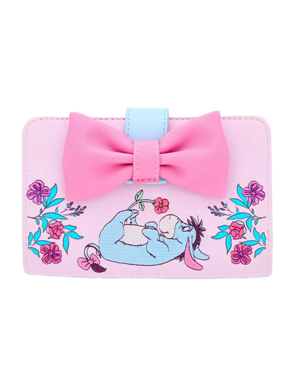 A pink wallet featuring a large pink bow on top, a light blue accent tab, and an embroidered illustration of Eeyore lying down surrounded by pink flowers and blue leaves, set against a plain white background.