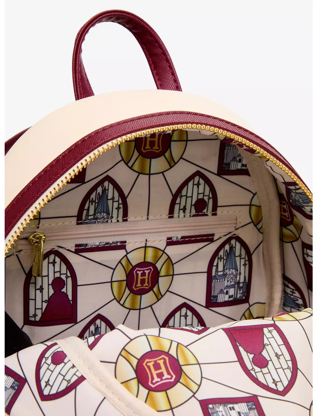 Close-up of a Hogwarts-themed backpack interior and exterior, featuring a cream-colored body with burgundy trim, gold zipper, and an all-over stained-glass window and crest pattern with castle motifs.