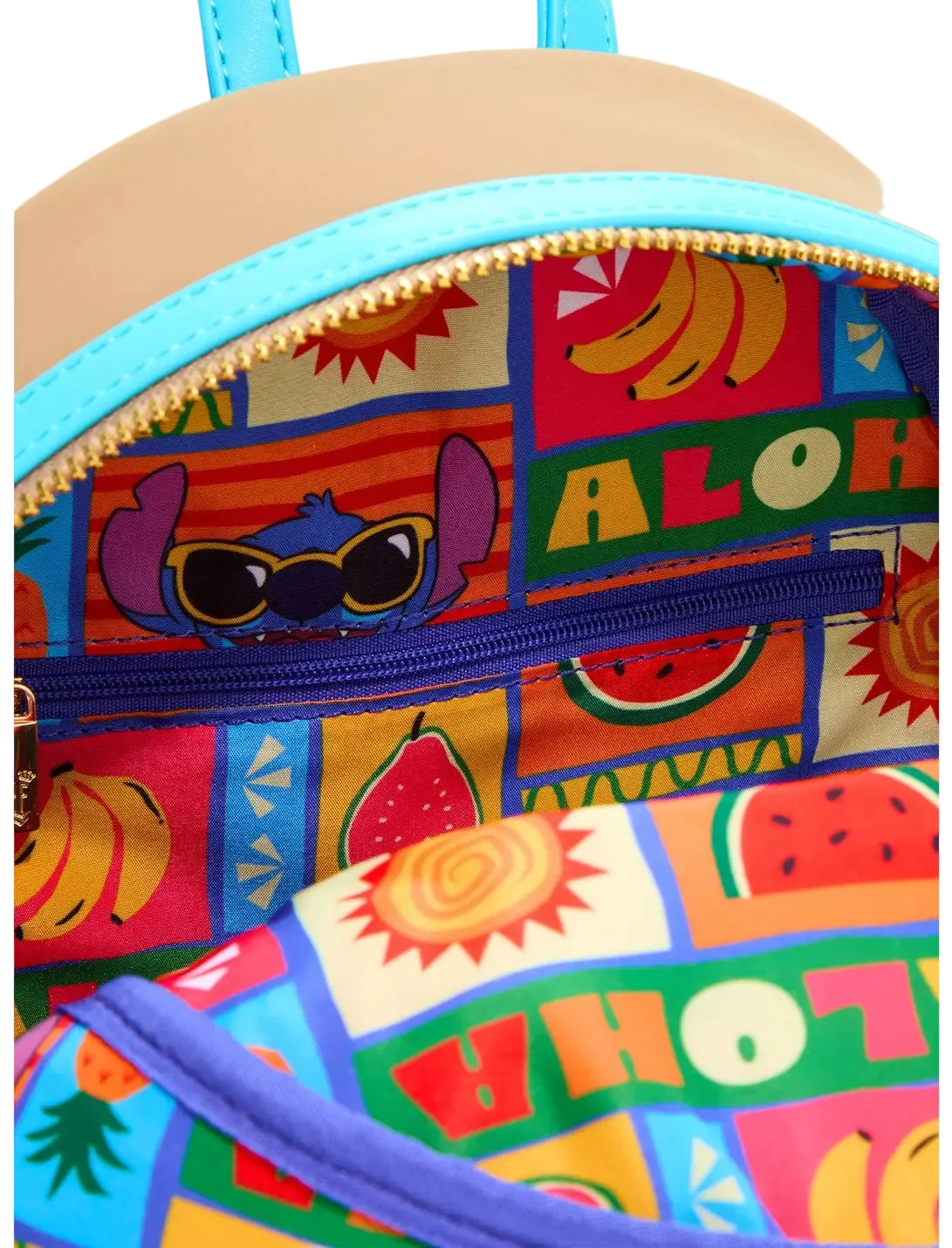 Close-up of a colorful backpack interior with a tropical Aloha patchwork print featuring cartoon characters, fruits, and text, with a turquoise zipper and a beige exterior visible at the top.
