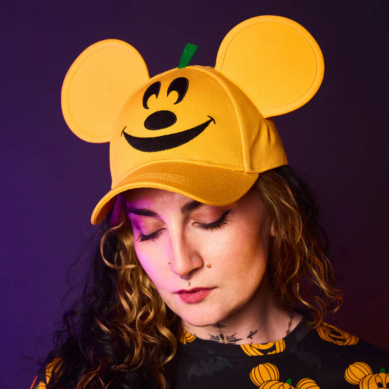 Person wearing a yellow cap with pumpkin ears and a smiley face against a purple background