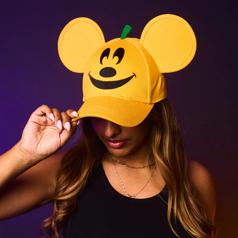 Person wearing a yellow cap with mouse ears and a pumpkin face design against a purple background