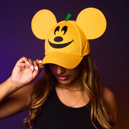 Person wearing a yellow cap with mouse ears and a pumpkin face design against a purple background