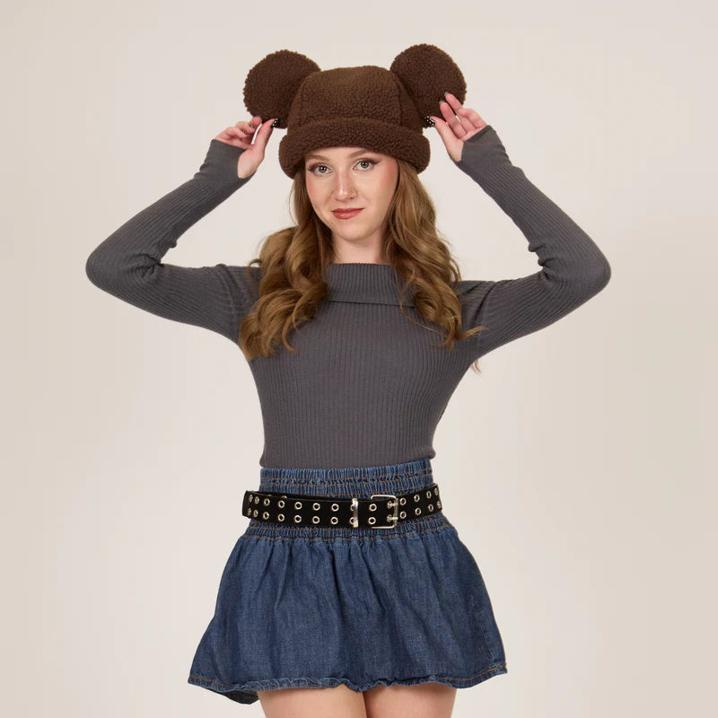 Woman wearing a brown bear hat with ears, gray sweater, and denim skirt on a beige background