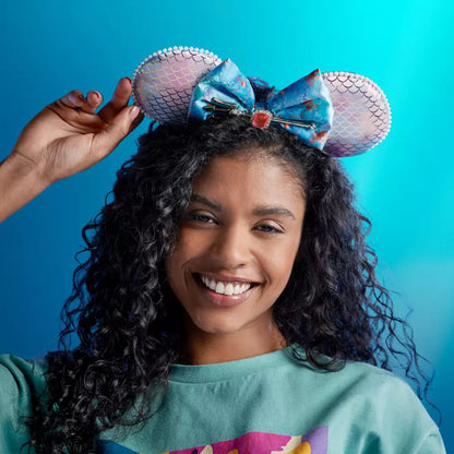 A smiling woman with curly dark hair wears a mermaid-themed headband with a blue patterned bow and mermaid-scale mouse ears against a bright blue background.