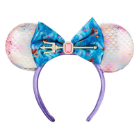 A mermaid-themed mouse ear headband featuring iridescent pink and white mermaid-scale ears, a large blue floral bow with a trident and pink gem accent, and a purple headband.