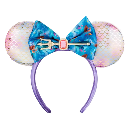 A mermaid-themed mouse ear headband featuring iridescent pink and white mermaid-scale ears, a large blue floral bow with a trident and pink gem accent, and a purple headband.