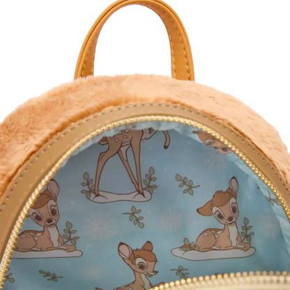 Close-up of a plush mini backpack with a light blue interior printed with Bambi the deer illustrations, tan faux-leather trim and handle, and a gold zipper.