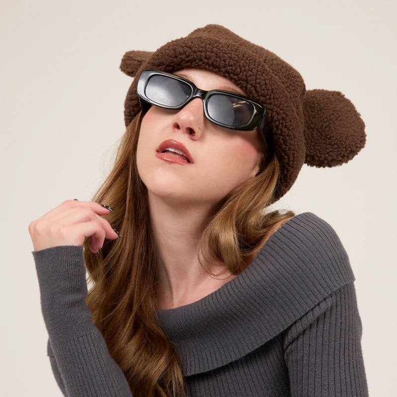 Woman wearing a brown knit hat with ears and dark sunglasses against a plain background