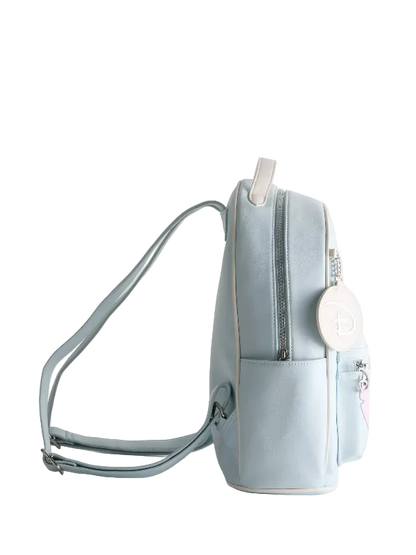 A light blue faux-leather backpack with a front pocket, silver zippers, and a white circular charm featuring a monogram logo, shown against a plain light gray background.