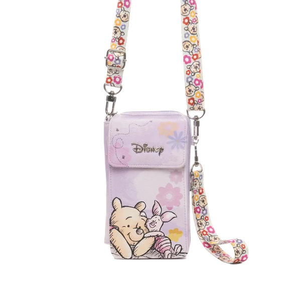 A Disney crossbody phone pouch in light purple, featuring Winnie the Pooh and Piglet illustrations, with a floral patterned strap and silver-tone hardware.