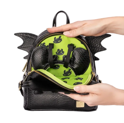 A black dragon-themed backpack with a textured python-skin style exterior, gold zippers, and wing-like side details, shown open with hands holding it to reveal a bright lime green interior lined with black dragon silhouettes.