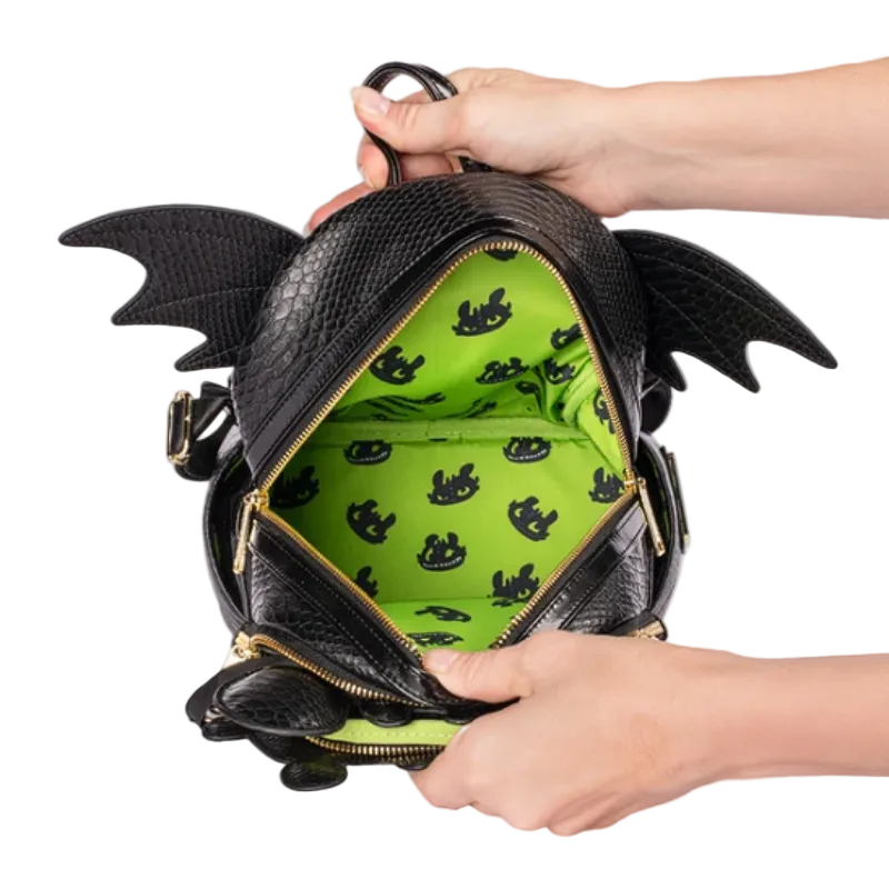 A black dragon-themed backpack with textured faux-leather finish, gold zippers, and a bright green interior patterned with small black dragon silhouettes. Hands are shown holding the open bag against a white background.