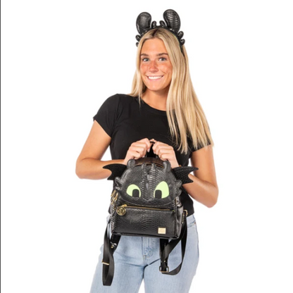 A woman wearing a black Toothless-themed headband holds a matching black faux-croc backpack shaped like Toothless from The Nightmare Before Christmas. The backpack features a black crocodile texture, gold zipper, and glowing green eyes, while the model wears a black t-shirt and light blue jeans against a solid white background.